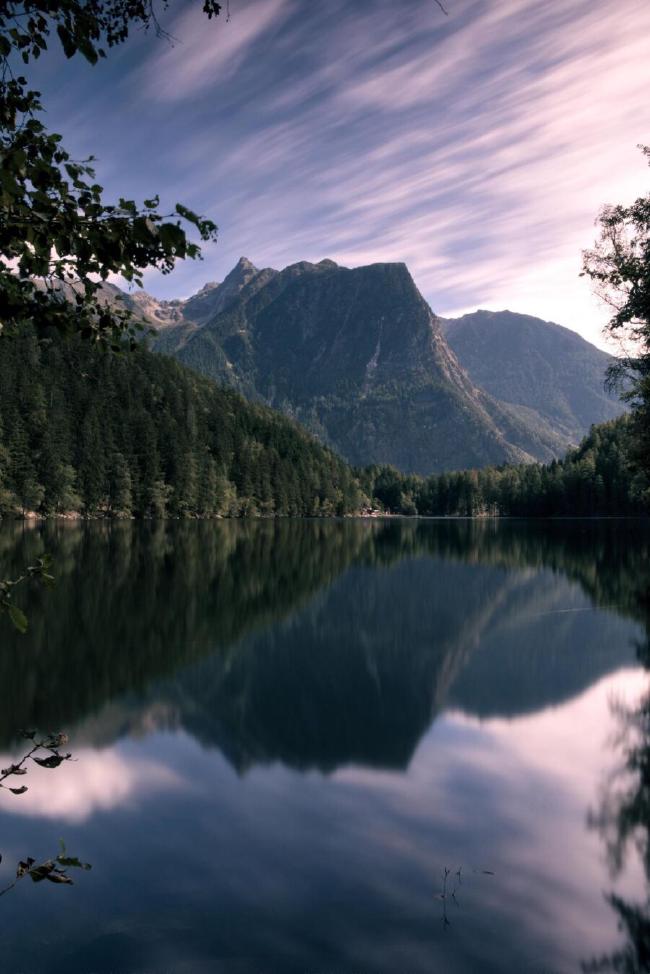 Longtime exposure of a lake with a beautiful mountain landscape behind it. It is sunny and there are only a few clouds on the blue sky. The clouds are motion blured, the light ist softer and you can see a reflection of the mountain in the sea